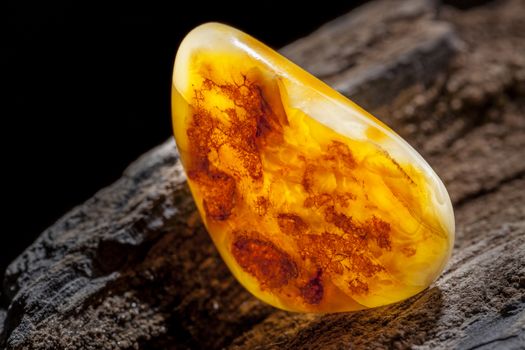 A piece of yellow semi opaque natural amber, classification color Bastard, has cracks inside. Polished, has a bead shape. Placed on dark stoned wood texture.