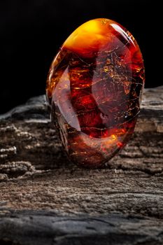 A piece of dark red semi transparent natural amber, classification color cherry or dragon blood, has cracks on its surface. Polished, oval shape. Placed on dark stoned wood texture.