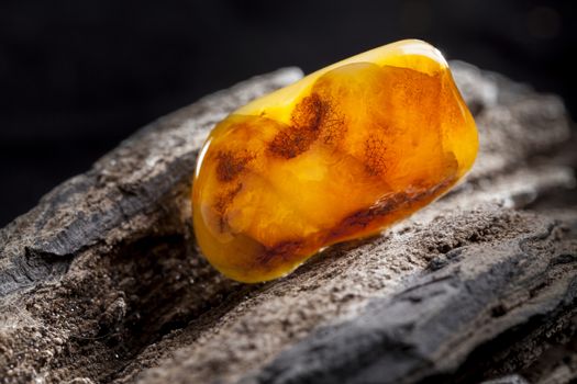 A piece of yellow semi opaque natural amber, classification color Bastard, has cracks inside. Polished, has a bead shape. Placed on dark stoned wood texture.