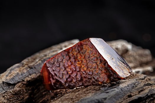 A piece of dark red semi transparent natural amber, classification color cherry or dragon blood, has cracks on its surface. Polished, oval shape. Placed on dark stoned wood texture.