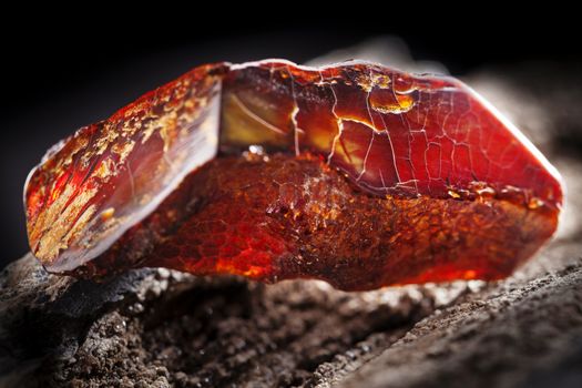 A piece of dark red semi transparent natural amber, classification color cherry or dragon blood, has cracks on its surface. Polished, oval shape. Placed on dark stoned wood texture.