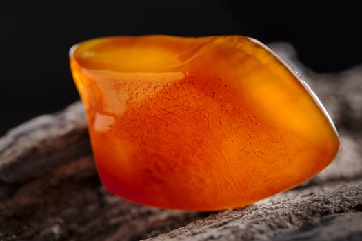 A piece of yellow semi transparent natural amber, classification color honey or cognac, has cracks on its surface. Polished, natural shape. Placed on dark stoned wood texture.