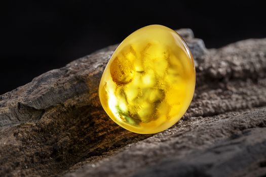 A piece of yellow semi opaque natural amber, classification color Bastard, has cracks inside. Polished, has a bead shape. Placed on dark stoned wood texture.