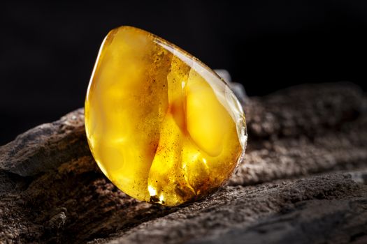 A piece of yellow semi opaque natural amber, classification color Bastard, has cracks inside. Polished, has a bead shape. Placed on dark stoned wood texture.