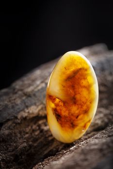 A piece of yellow semi opaque natural amber, classification color Bastard, has cracks inside. Polished, has a bead shape. Placed on dark stoned wood texture.