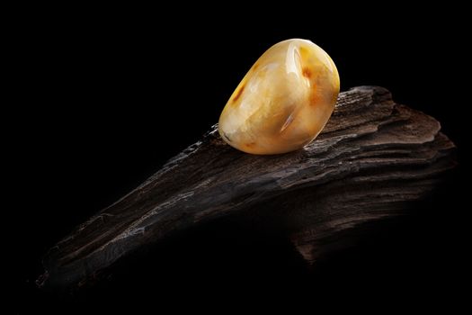 A piece of yellow semi opaque natural amber, classification color Bastard, has cracks inside. Polished, has a bead shape. Placed on dark stoned wood texture.