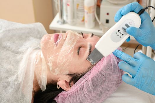 Young woman in beauty salon doing peeling and facial cleansing procedure. Cosmetic multifunctional device. Ultrasound procedure. Medical equipment healthcare. Face professional massage.