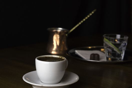 a glass of coffee with coffee pot and chocolate beside a coffee with milk in a white cup placed on a brown table