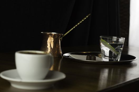 a glass of coffee with coffee pot and chocolate beside a coffee with milk in a white cup placed on a brown table