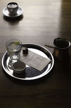 a glass of coffee with coffee pot and chocolate beside a coffee with milk in a white cup placed on a brown table