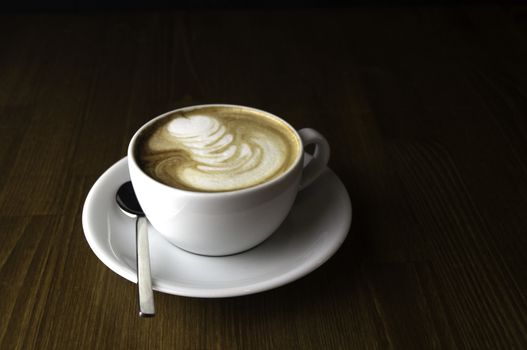 coffee with milk in a white cup on a brown table and spoon to mix