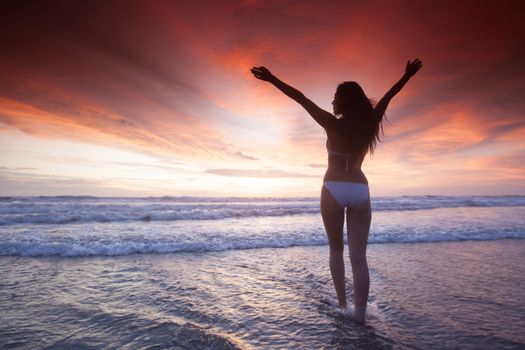 Woman in bikini raising arms towards beautiful glowing sunset over sea