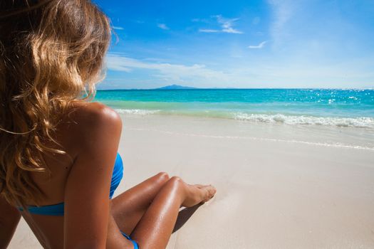 Woman on beach sitting in sand looking at ocean enjoying sun and summer travel holidays vacation getaway. Girl in bikini relaxing under blue sky.