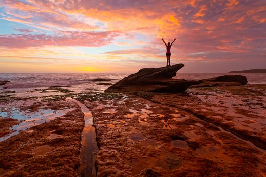 Female standing on top of Shark rock on the coastal rock shelf admiring the magnificent sunrise which is also reflected in the coatal rock shelf and tidal waters.