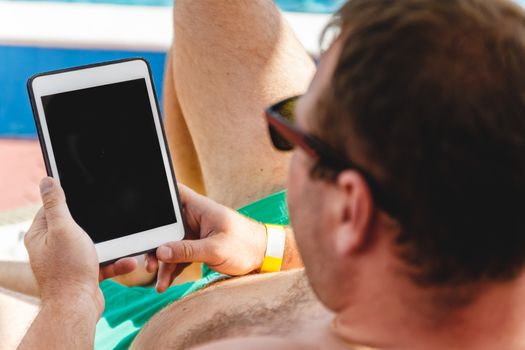 A young man in sunglasses holds a tablet in his hands. Freelancer job concept on the beach and vacation.