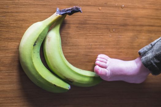 Bare foot touching a pair of green bananas on a wooden table