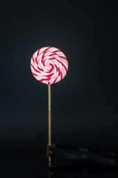 Colorful lolipop isolated on plain black background, wooden stick held bz clamp, red, rose and white spiral, childhood sweets, also unhealthy junk food concept