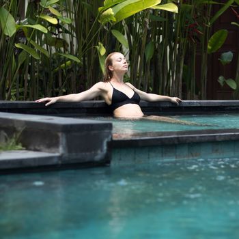 Sensual young woman relaxing in outdoor spa infinity swimming pool surrounded with lush tropical greenery of Ubud, Bali. Wellness, natural beauty and body care concept.