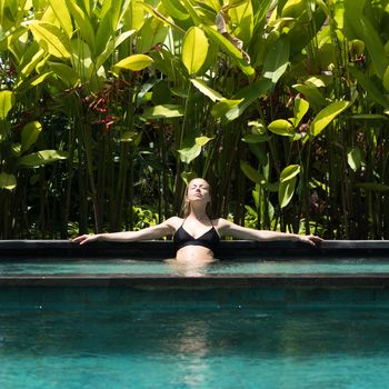 Sensual young woman relaxing in outdoor spa infinity swimming pool surrounded with lush tropical greenery of Ubud, Bali. Wellness, natural beauty and body care concept.