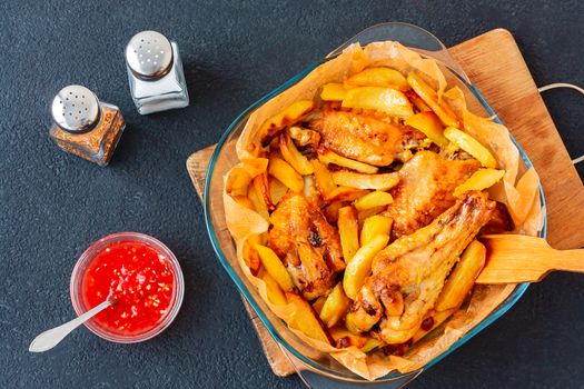 Baked turkey wings with potato pieces in a square baking dish on a black kitchen table with sauce and spices - photo, image.