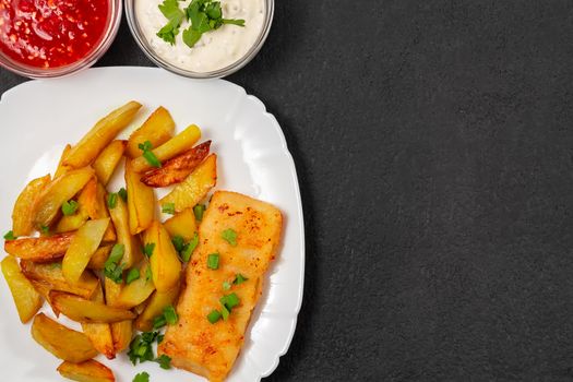 Fried fish and chips on a white plate on the kitchen table with tomato sauce, tartar sauce and pickled vegetables salad - photo, image. Flat lay, top view.