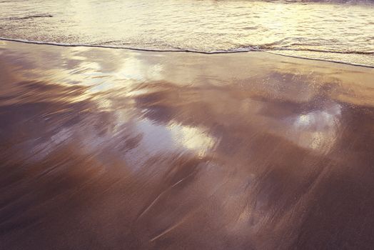 soft wave on the sand of the seashore at sunset, the water on the surface reflects the sky and the sunlight