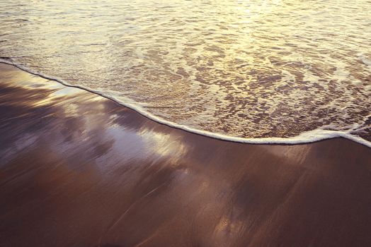 soft wave on the sand of the seashore at sunset, the water on the surface reflects the sky and the sunlight