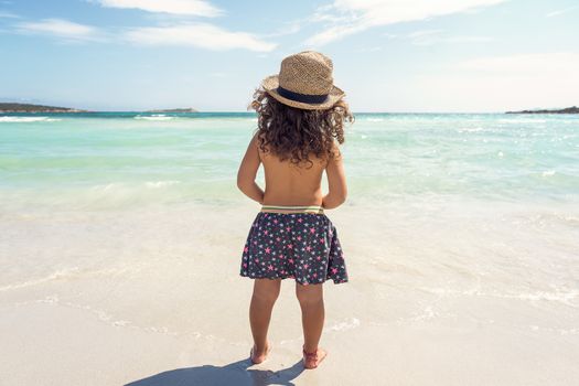 little girl with her back on the shore of a paradisiacal beach of white sand and turquoise waters, wearing a skirt and a cane hat