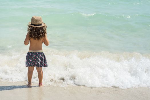 little girl with her back on the shore of a paradisiacal beach of white sand and turquoise waters, wearing a skirt and a cane hat