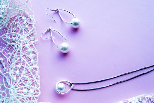 Beautiful silver shiny pearl jewelry with trendy neon glow, trendy glamorous earrings, chain on pink purple background with exquisite lace. Flat lay, top view, copy location