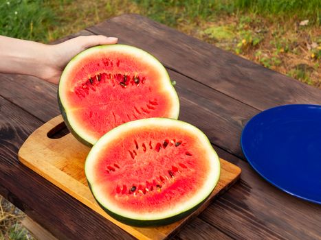 Female hands cut a ripe watermelon on a wooden table with a knife.