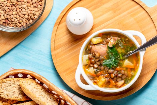 Lentil soup with chicken in a white bowl on a wooden board on a blue table, top view.