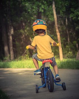 Vintage tone rear view of Asian toddler boy riding tricycle at nature park near Dallas, Texas, USA. Healthy kid cycling with helmet, short and sneaker on park concrete pathway