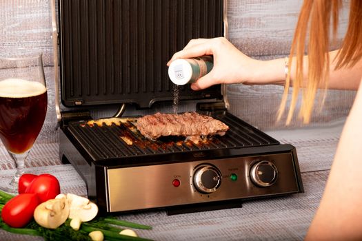 Beef is cooked on the electric grill. Around the grill vegetables laid out. On a wooden background.