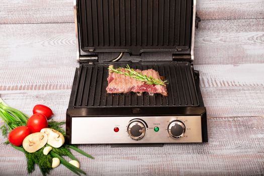 Beef is cooked on the electric grill. Around the grill vegetables laid out. On a wooden background.