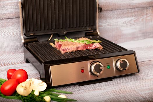 Beef is cooked on the electric grill. Around the grill vegetables laid out. On a wooden background.