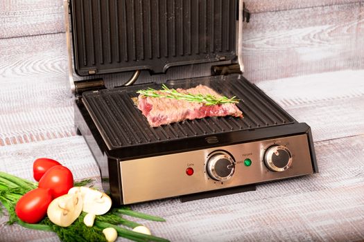 Beef is cooked on the electric grill. Around the grill vegetables laid out. On a wooden background.