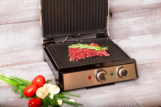 Beef is cooked on the electric grill. Around the grill vegetables laid out. On a wooden background.