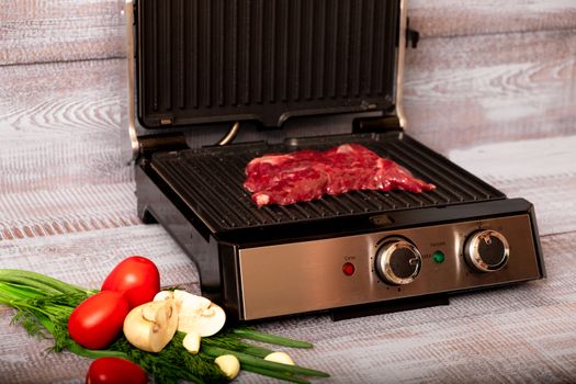 Beef is cooked on the electric grill. Around the grill vegetables laid out. On a wooden background.