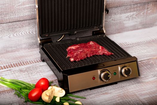 Beef is cooked on the electric grill. Around the grill vegetables laid out. On a wooden background.