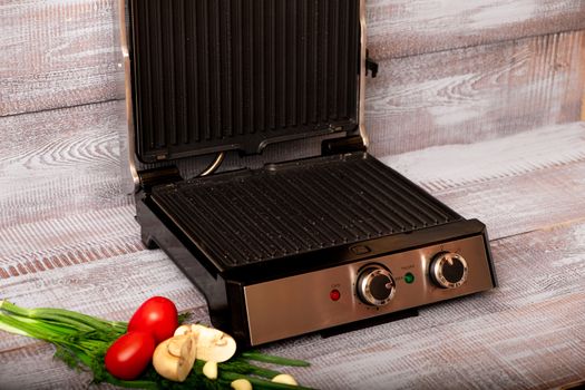 Beef is cooked on the electric grill. Around the grill vegetables laid out. On a wooden background.