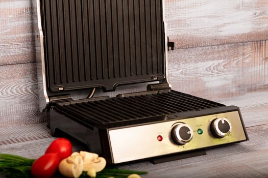 Beef is cooked on the electric grill. Around the grill vegetables laid out. On a wooden background.