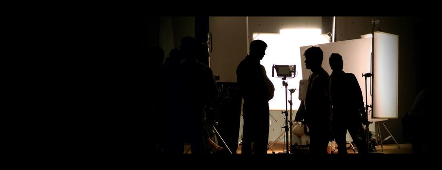 Shooting studio behind the scenes in silhouette images which film crew team working for filming movie or video with professional lighting and equipment such as camera, tripod, soft box, monitor