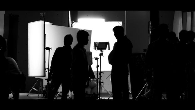 Shooting studio behind the scenes in silhouette images which film crew team working for filming movie or video with professional lighting and equipment such as camera, tripod, soft box, monitor