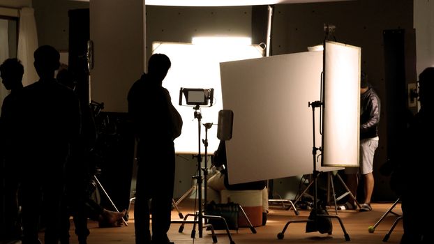 Shooting studio behind the scenes in silhouette images which film crew team working for filming movie or video with professional lighting and equipment such as camera, tripod, soft box, monitor