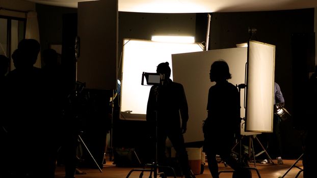 Shooting studio behind the scenes in silhouette images which film crew team working for filming movie or video with professional lighting and equipment such as camera, tripod, soft box, monitor