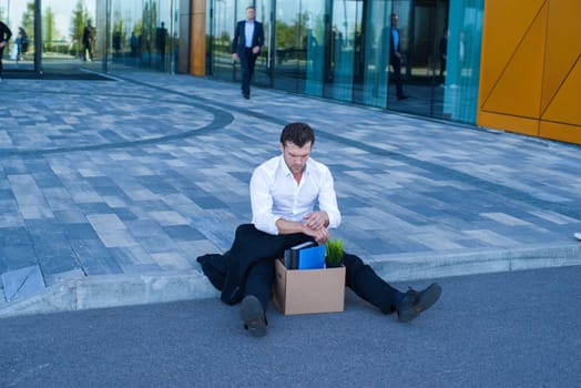 Fired business man sitting frustrated and upset on the street near office building with box of his belongings. He lost work