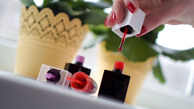 Pair of female hands opening nail polish bottle. Nail polishes on the bright window-sill with flowers