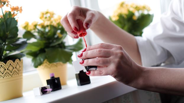 Pair of female hands opening nail polish bottle. Nail polishes on the bright window-sill with flowers