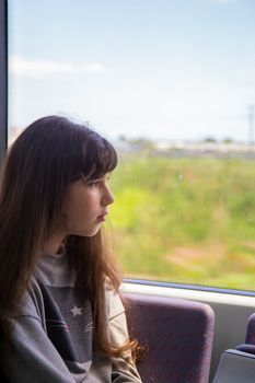 Beautiful teenage girl with long brown hair rides a commuter train and looks out the window.